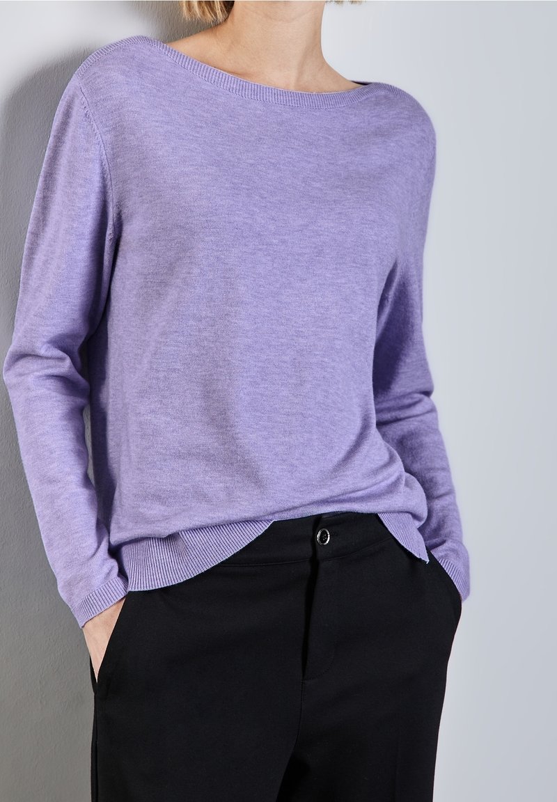 Person wearing a lavender long-sleeve sweater tucked into black pants with hands in pockets, standing against a light gray wall.