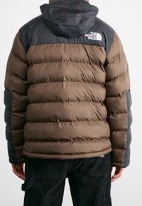 Chaqueta acolchada marrón con hombros y capucha negros, con patrón acolchado y el logo de The North Face. El material parece liso y aislante.