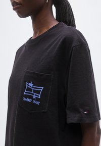 Čierne bavlnené tričko s krátkym rukávom, s modrým vyšívaným vzorom na vrecku, ktorý obsahuje text "TOMMY JEANS" a tenké prešívanie.
