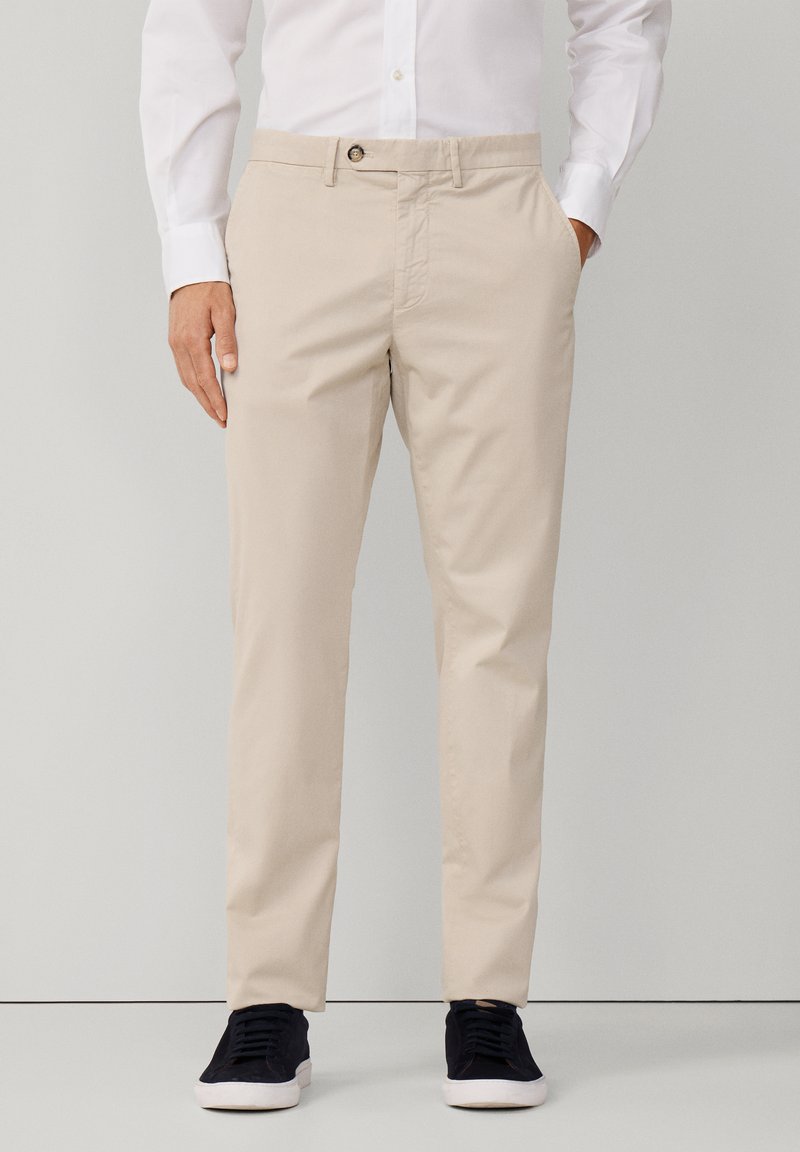 Chino beige con frontale piatto e tasche laterali, dotati di chiusura a bottone. Abbinati a sneakers scure, che valorizzano il look casual.