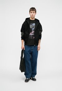 Jauns vīrietis stāv, ģērbies melnā kapucē ar baltām tīkla dizaina detaļām uz piedurknēm, grafisko t-kreklu, brīvām zilām biksēm, melnām sporta apavām un tur melnu somu.