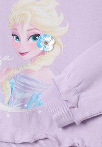 Lavender sweatshirt featuring a printed character design, embellished with rhinestones and a floral accent. Ruffled cuffs and hem detail.