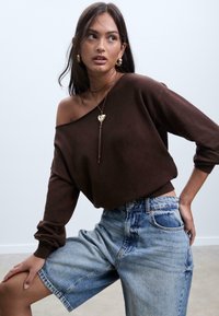 Brauner schulterfreier Pullover aus weichem Material, kombiniert mit hellblauen Denim-Shorts. Das Modell trägt goldene Ohrringe und eine Anhänger-Halskette.