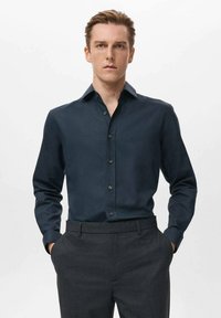 Camisa de manga larga azul oscuro hecha de tela suave, con cuello puntiagudo y cierre frontal de botones, combinada con pantalones grises de corte sastre.