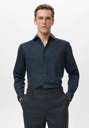 Camisa de manga larga azul oscuro hecha de tela suave, con cuello puntiagudo y cierre frontal de botones, combinada con pantalones grises de corte sastre.