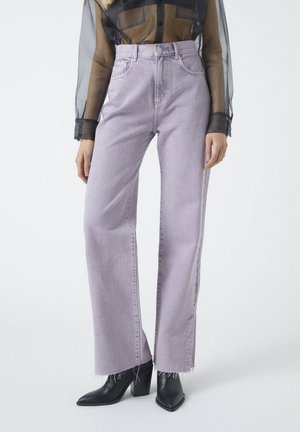 Person trägt hoch taillierte, hellviolette, weit geschnittene Jeans mit ausgefransten Säumen, schwarze spitze Stiefel und ein durchsichtiges schwarzes Langarmshirt.