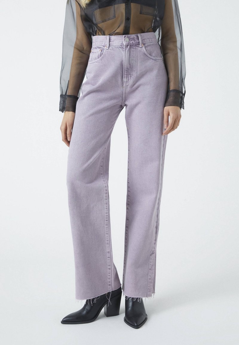 Personne portant un jean large taille haute violet clair avec des ourlets effilochés, des bottes noires pointues et un haut noir transparent à manches longues.