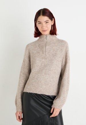 Beige kötött pulóver magas gallérral és félcipzárral, amely puha, texturált felülettel rendelkezik. Fekete bőrszoknya kíséretében.