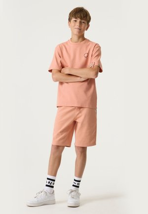 Garçon debout, bras croisés, portant un t-shirt pêche et un short, des baskets blanches et des chaussettes à motifs noir et blanc, sur un fond uni.