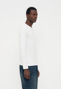 Άνδρας που φοράει απλό λευκό μακρυμάνικο πουκάμισο και μπλε τζιν, στέκεται πλαγίως μπροστά σε ένα απλό λευκό φόντο.