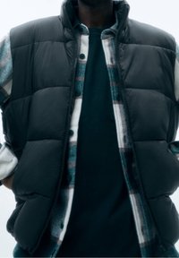 Gilet imbottito nero sopra una camicia a scacchi verde e bianca, indossato con un maglione scuro. Presenta sezioni imbottite e un colletto alto.