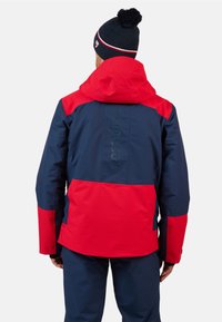 Giacca impermeabile rossa e blu navy con cappuccio, pannelli a contrasto e tessuto esterno testurizzato. Presenta un dettaglio con logo sul retro.