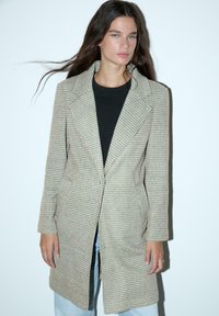 Bež plašč s houndstooth vzorcem in prilagojeno zasnovo, z velikimi ovratniki in stranskimi žepi, v kombinaciji s črnim topom in kavbojskimi hlačami.