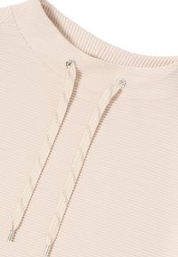 Beige rippad hoodie med hög krage, prydd med två gräddfärgade snören med silvermetallspetsar, som visar upp ett mjukt, strukturerat tyg.