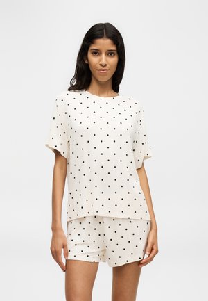 Woman wearing a white short-sleeve top and matching shorts with black polka dots, standing against a plain background.
