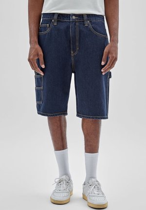 Mężczyzna w ciemnoniebieskich krótkich spodniach typu cargo z denimu, białych skarpetkach sportowych i białych trampkach, stojący na jednolitym tle.