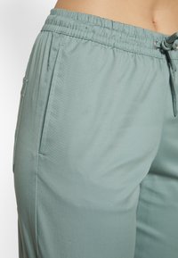 Calças de jogging verde clara feitas de um tecido suave, apresentando uma cintura elástica com cordão e bolsos laterais. Design simples e ajustado.