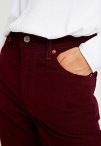 Pantalon en velours côtelé bordeaux à coupe ajustée, avec des poches standard et une fermeture à bouton métallique. Tissu texturé à côtes verticales.