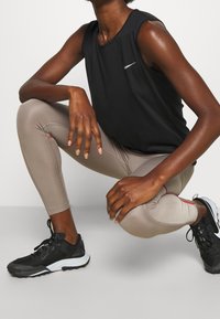Musta hihaton urheilu paita, beige leggings punaisilla yksityiskohdilla, mustat lenkkitossut. Malli kyykistyy, esitellen urheiluvaatteiden yksityiskohtia ja istuvuutta.