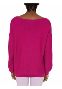 Vue arrière d'une personne portant un pull ample et lumineux de couleur magenta avec des manches longues et un col bateau, associé à un pantalon de couleur claire.