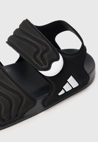 Μαύρο αθλητικό σανδάλι με ρυθμιζόμενους επενδεδυμένους ιμάντες και λευκές λεπτομέρειες λογότυπου στη σόλα με υφή, εμφανιζόμενο σε κοντινή γωνία σε λευκό φόντο.