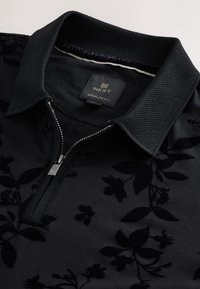Polo noir avec col texturé et motif floral, doté d'une fermeture éclair argentée partiellement ouverte et d'une étiquette indiquant "NEXT Regular Fit."