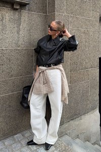 Chemise en cuir noir surdimensionnée, pantalon blanc ample, écharpe beigne à carreaux, ceinture noire, sac noir, chaussures noires à bout pointu, lunettes de soleil. Fond de mur texturé.