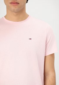 Ljusrosa bomulls-T-shirt med korta ärmar och rund halsringning, med en liten broderad logotyp nära bröstet i röd, vit och blå.