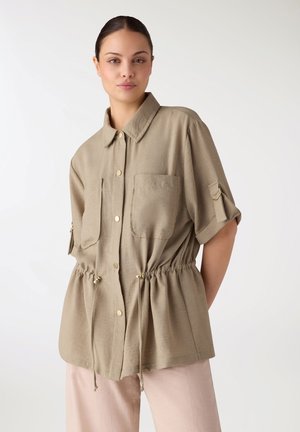 Donna indossa una camicia beige con bottoni frontali, tasche e coulisse in vita, abbinata a pantaloni rosa chiaro, in piedi davanti a uno sfondo bianco.