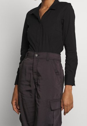 Femme portant une chemise noire à col à manches longues rentrée dans un pantalon cargo sombre taille haute avec une poche latérale à rabat.