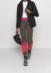 Czarna wodoodporna kurtka z kapturem, w parze z brązowymi spodniami z różowymi akcentami. Strój uzupełniają szare buty trekkingowe i kask.