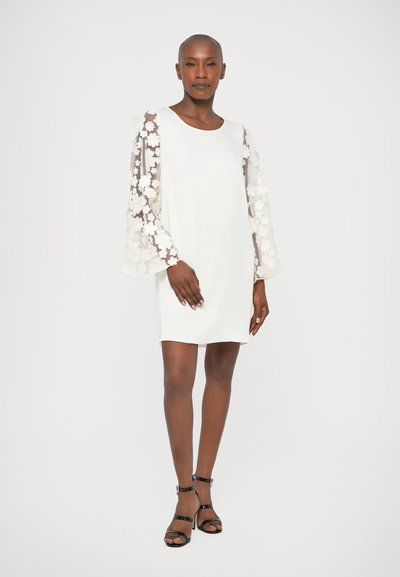 Vestido mini blanco con mangas de campana de encaje floral, escote redondo y textura suave. Llevado con tacones negros con tiras sobre un fondo neutro.