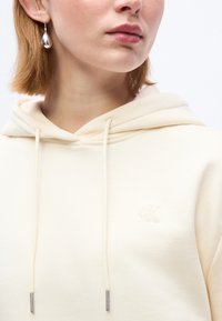 Κοντινό πλάνο ενός ατόμου που φοράει κρεμ φούτερ με κουκούλα και λογότυπο cK, και ασημένιο σκουλαρίκι σε σχήμα σταγόνας, εμφανίζοντας το λαιμό, τα χείλη και μέρος του πηγουνιού και των μαλλιών.