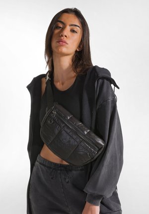 Borsa a tracolla nera in nylon con superficie testurizzata, numerose tasche con zip e tracolla regolabile. Indossata sopra un outfit nero.