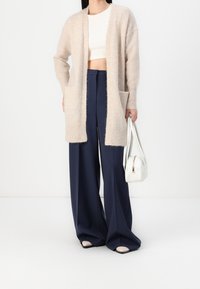 Béžový pletený kardigan s otevřeným předním dílem, dvěma předními kapsami a dlouhými rukávy, doplněný tmavě modrými širokými kalhotami a bílou kabelkou.