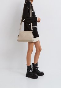 Beige kožená kabelka přes rameno s vyraženým chevronovým vzorem, stříbrnými kováními a odnímatelným popruhem. Kontrastní stehy podtrhují design.