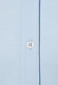 Camicia azzurra chiara con tessuto texturizzato, dettaglio dei bottoni con design a quattro fori e cuciture visibili lungo la profilo.