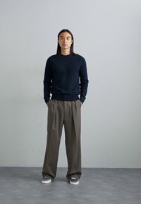 Model trägt einen dunkelblauen Strickpullover und hellbraune, gefaltete Hosen. Die Schuhe sind grau mit weißen Sohlen. Der Hintergrund ist hellgrau.