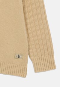 Beige strikkegenser med ribbestrikkede mansjetter og hem, med et distinkt merkemerke med initialene 'cK' i en rektangulær lapp.