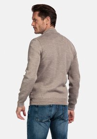 Man die met zijn rug naar de camera staat, gekleed in een beige lange mouwen trui en een blauwe spijkerbroek, tegen een effen witte achtergrond.