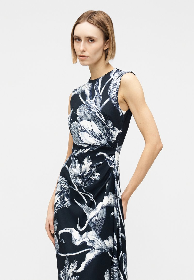 Woman with short hair wearing a sleeveless black dress with large white floral print, standing with one hand on her hip.