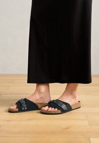 Personne portant des sandales noires à enfiler avec des semelles en liège et une longue jupe noire, debout sur un sol en bois clair.