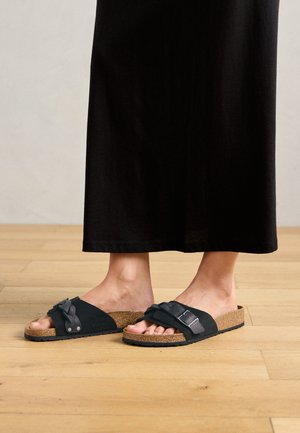 Person wearing black slip-on sandals with cork soles and black long skirt, standing on light wooden floor.