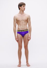 Lila férfi alsónadrág fekete derékpánttal, melyen sárga felirattal szerepel a "DSQUARED2". Az anyag sima és testhez álló, alacsony derékvonallal.
