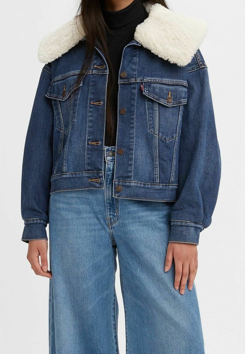 Farkkutakki, jossa pehmeä valkoinen sherpa-kaulus, edessä napillinen kiinnitys, rintataskut ja pitkät hihat, yhdistetty vaaleansinisiin, leveälahkeisiin farkkuihin.