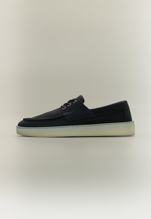 Melnas ādas ikdienas laivu kurpes ar baltu gumijas zolīti un priekšējo auklu, attēlotas sānskatā uz neitrālas krāsas fona.