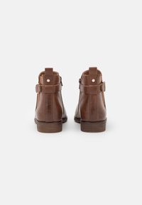 Bottines en cuir marron avec une texture lisse, présentant un détail de sangle et de boucle, et un petit talon empilé. Coutures visibles sur les côtés.