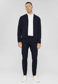 Navy cardigan, white shirt, and dark trousers. Smooth fabric, relaxed fit, and minimalistic design. Footwear: white sneakers.