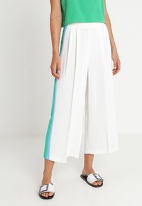 Pantalon blanc à jambes larges avec des bandes latérales vertes et bleues, présentant des plis et une texture lisse. Associé à des sandales blanches à enfiler.