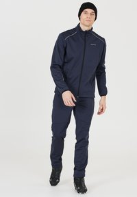 Marineblauwe jas en broek van rekbaar materiaal, met een ritssluiting aan de voorkant, zijzakken en een getailleerd ontwerp. Draagt met zwarte schoenen en een muts.